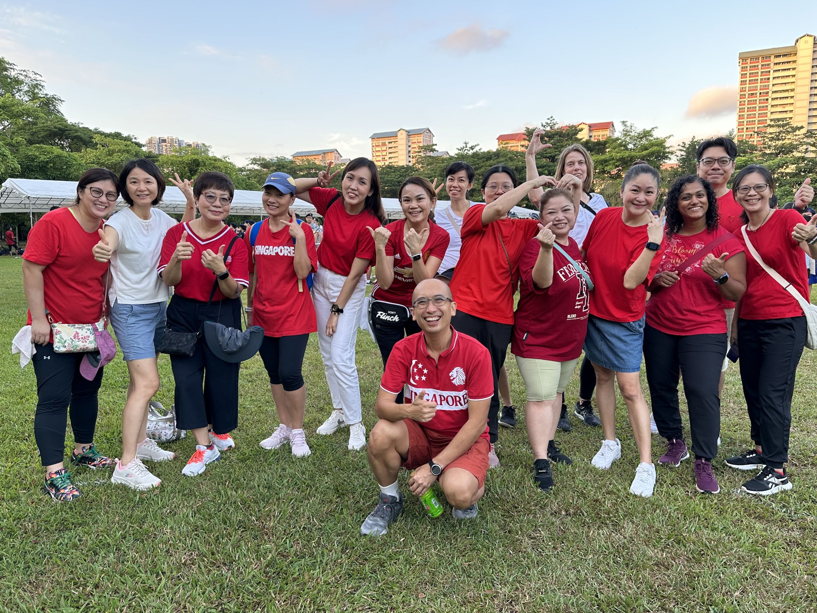 A large group of smiling people mostly in red Singapore shirts pose outdoors on grass with buildings and trees behind them.